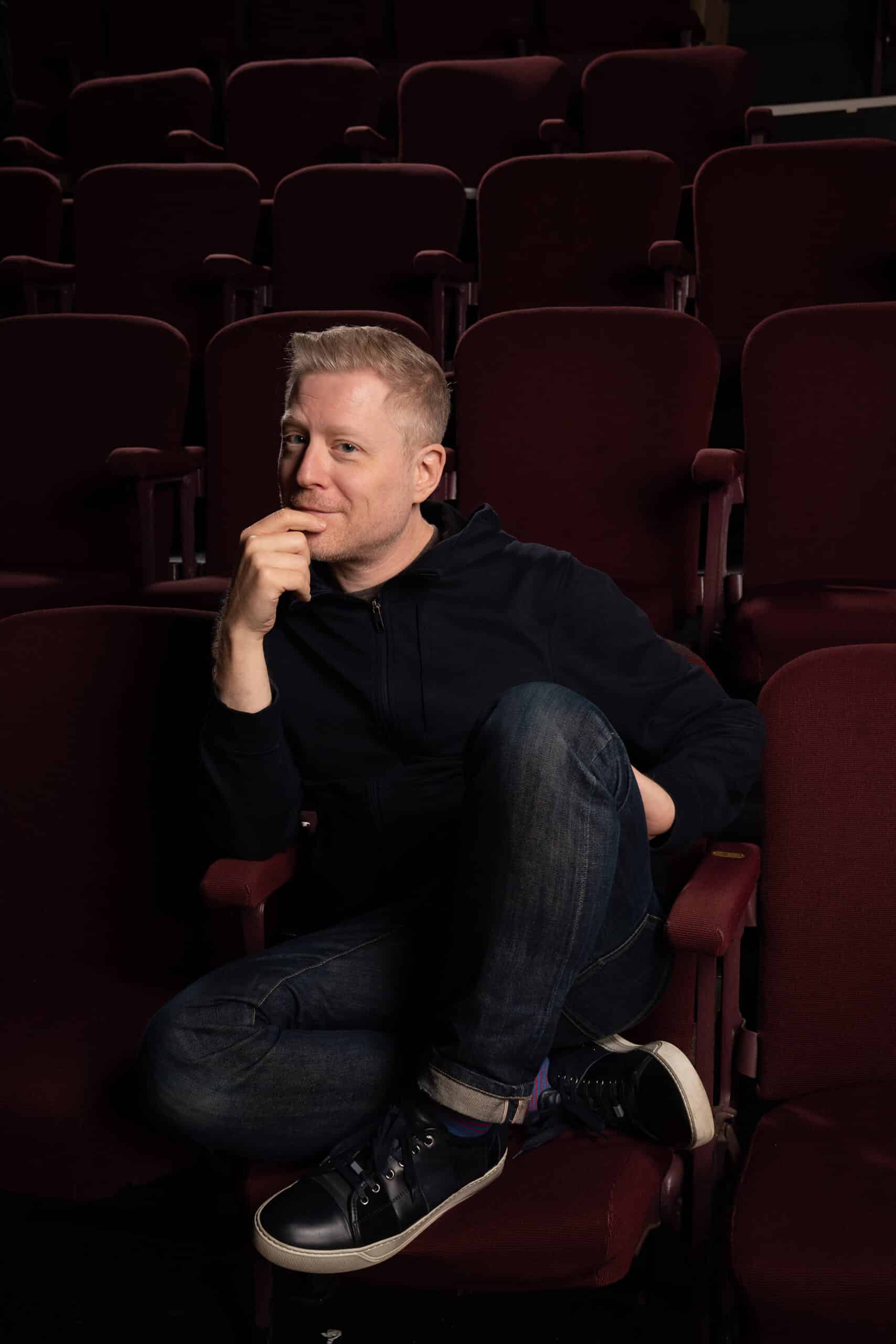 Anthony Rapp sitting in a theatre seat with his hand on his chin and a grin on his face.
